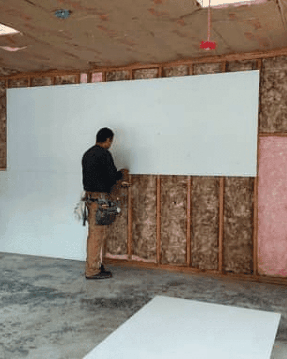 A member of the crew fitting a sheet of drywall over insulated stud framing, the ceiling joists still open above.